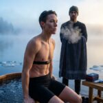 Athlete practicing Wim Hof breathing next to an outdoor ice plunge at dawn with a coach nearby, visible breath vapor and safety gear present