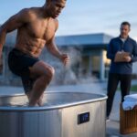 Athlete entering a cold plunge tub outdoors at dawn, digital thermometer reads 10–12°C, coach observing, towels nearby